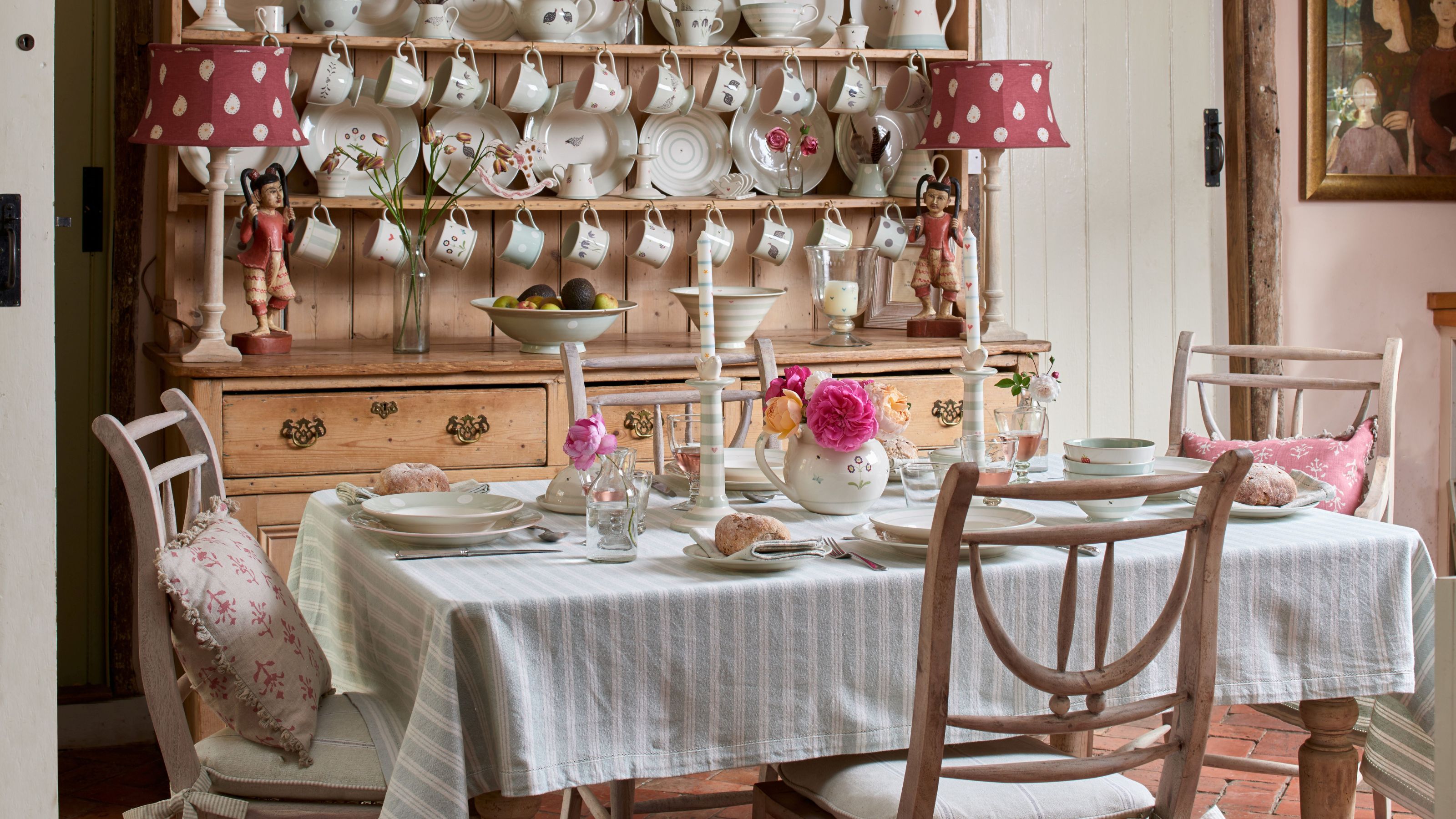 A dining table with a light-blue striped tablecloth and pink peonies, backed by a large wooden dresser with red patterned lamps