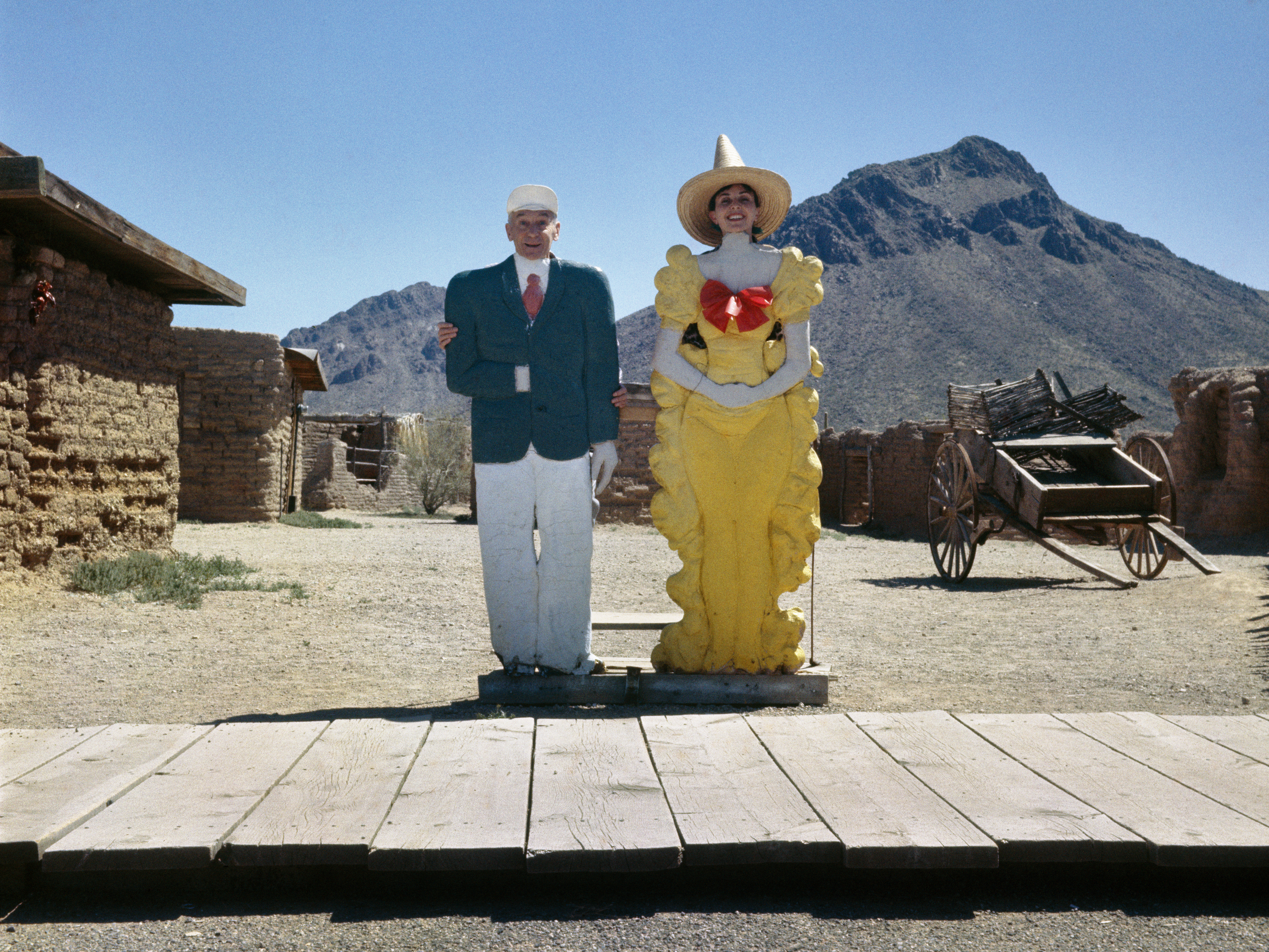 Two painted wooden cut-out figures, a man in a suit and a woman in a yellow dress with a sombrero, stand on a boardwalk against a desert mountain backdrop.