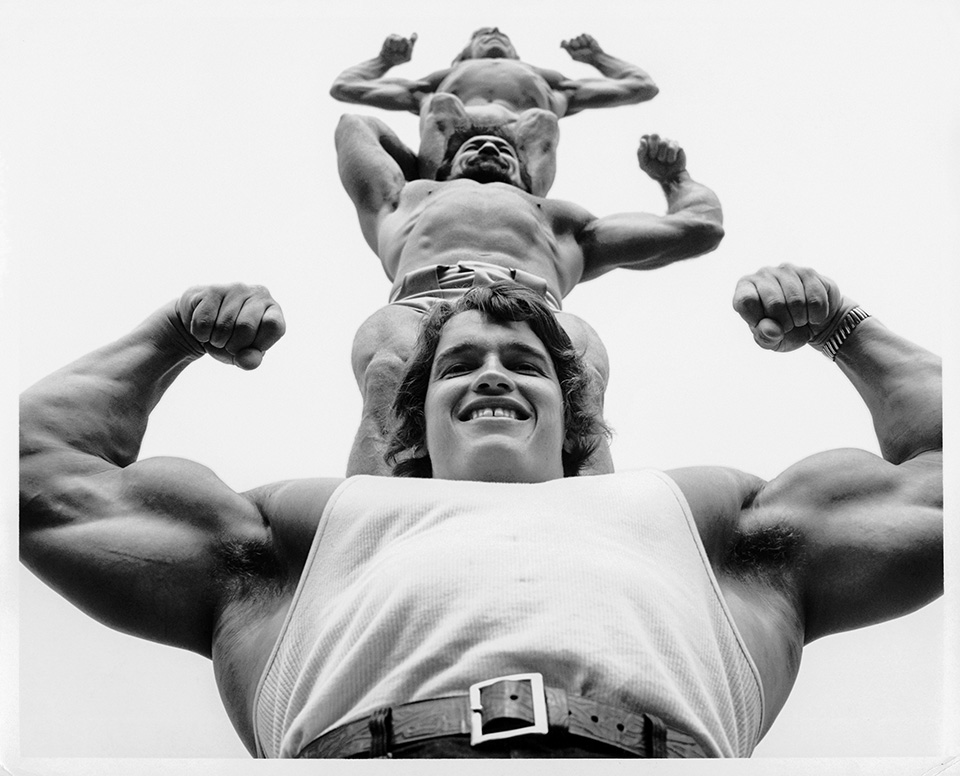 A low-angle black-and-white shot looking up at a grinning young Schwarzenegger flexing both biceps in a vest, with a second bodybuilder stacked on his shoulders doing the same above him.