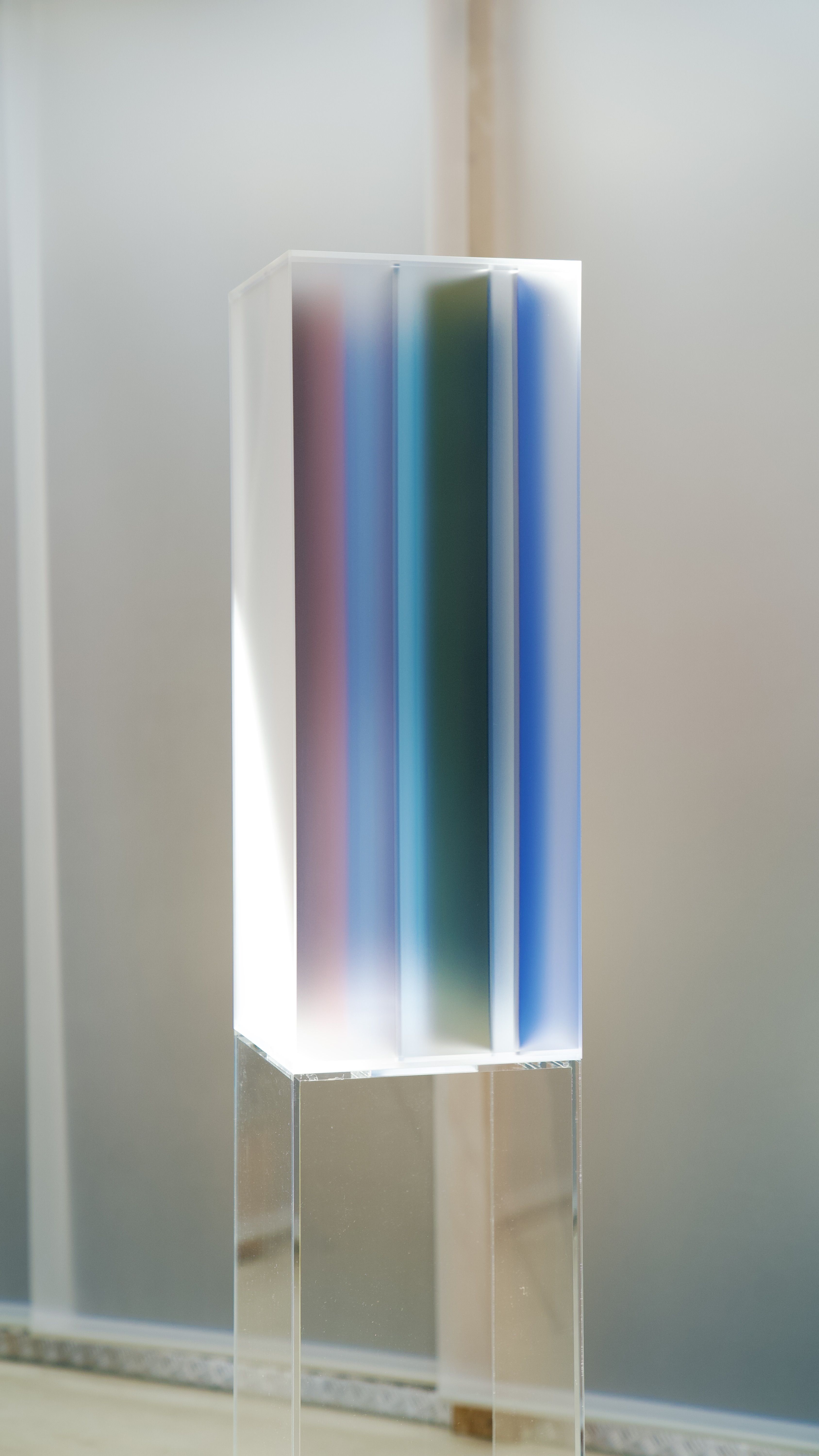 Installation view of an exhibition featuring gradient sculptural pieces made of opaque glass in different tones, placed atop transparent pedestals.