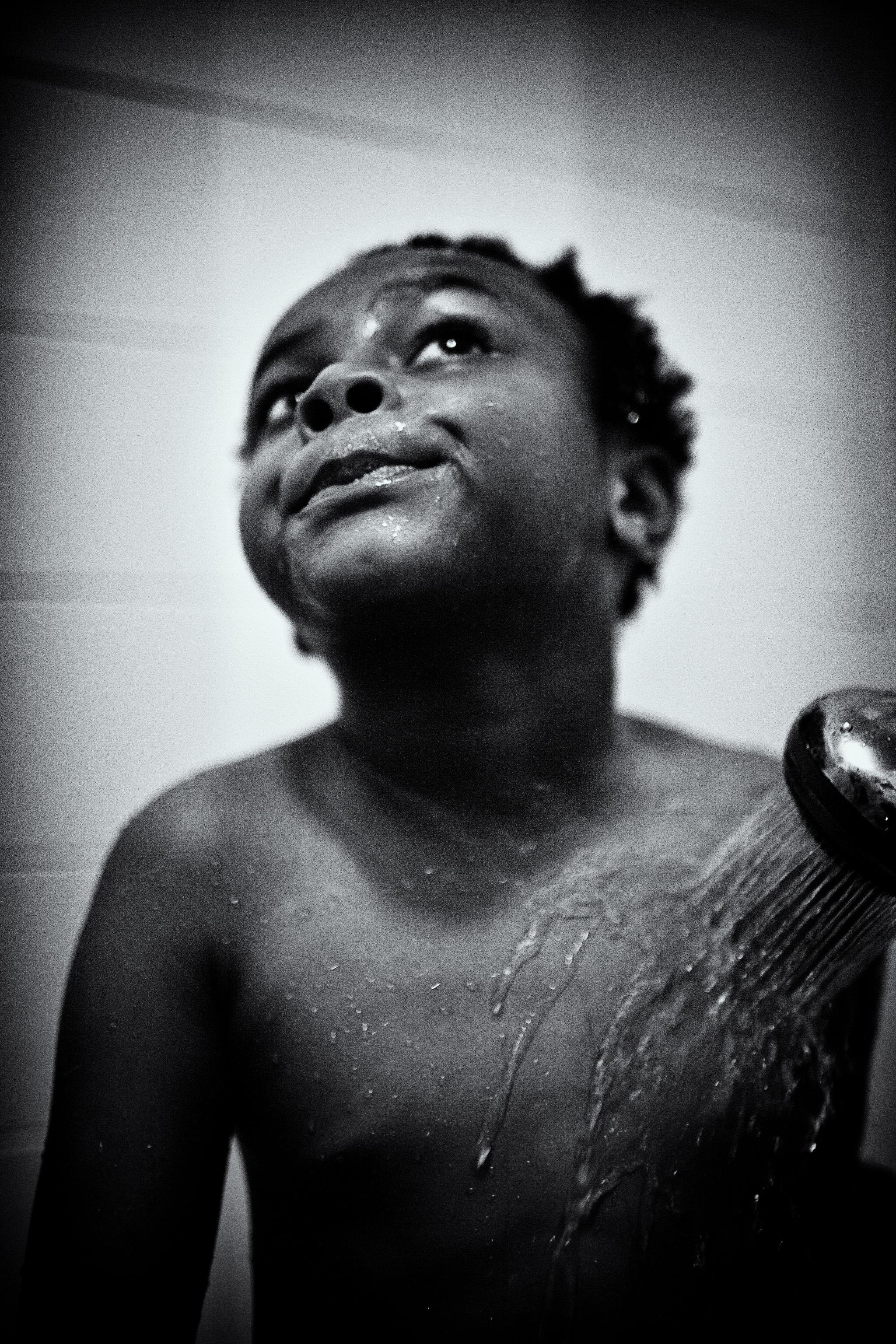 A black and white close-up photo of a young boy smiling and looking upward while water from a shower head rinses his shoulder.