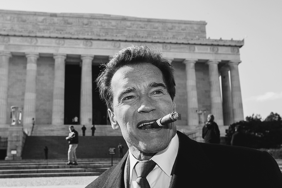 A square-format black-and-white portrait of Schwarzenegger in a Harley-Davidson cap and vest, staring directly into the camera while smoke billows from a cigar clamped between his lips.