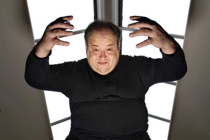 Rob Grant wearing a black jumper and looking down at the camera with his hands held up either side of his head, and bright lighting behind him.