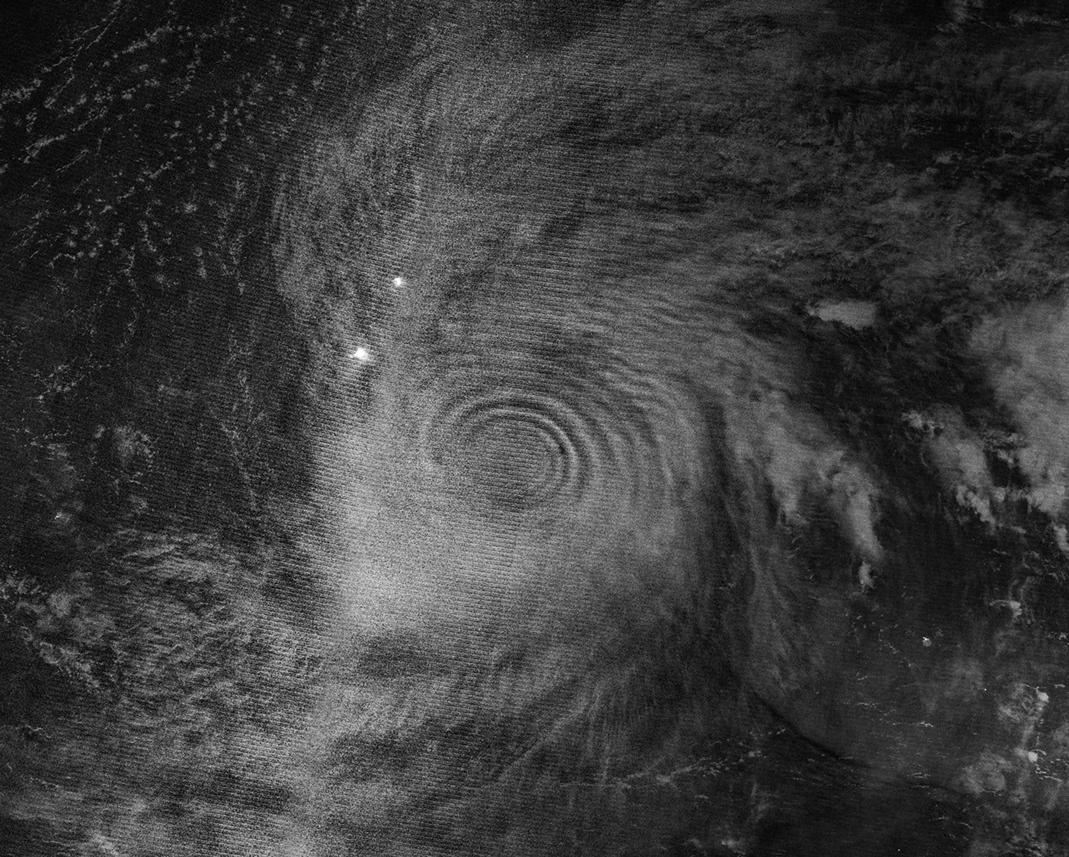 Gravity waves in the upper atmosphere appear as concentric rings in a nighttime, black and white satellite image. Clouds from a typhoon are also visible.