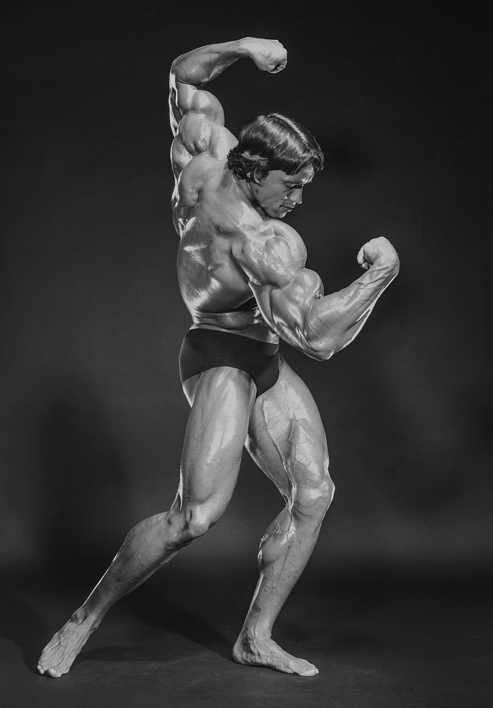 A black-and-white studio shot of Schwarzenegger performing a back double bicep pose, his oiled musculature catching dramatic side lighting against a dark grey background.