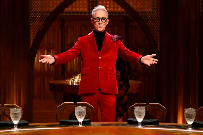 Alan Cumming wearing a red suit, stood behind the round table with his arms stretched outward.