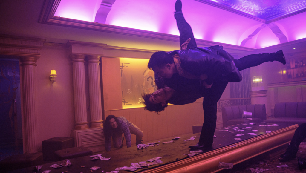 A man in a suit performs a mid-air martial arts kick against another man in a neon-lit room with scattered cash.