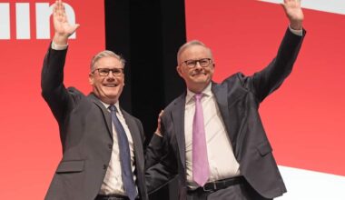 Two men in suits waving on stage against a red background.