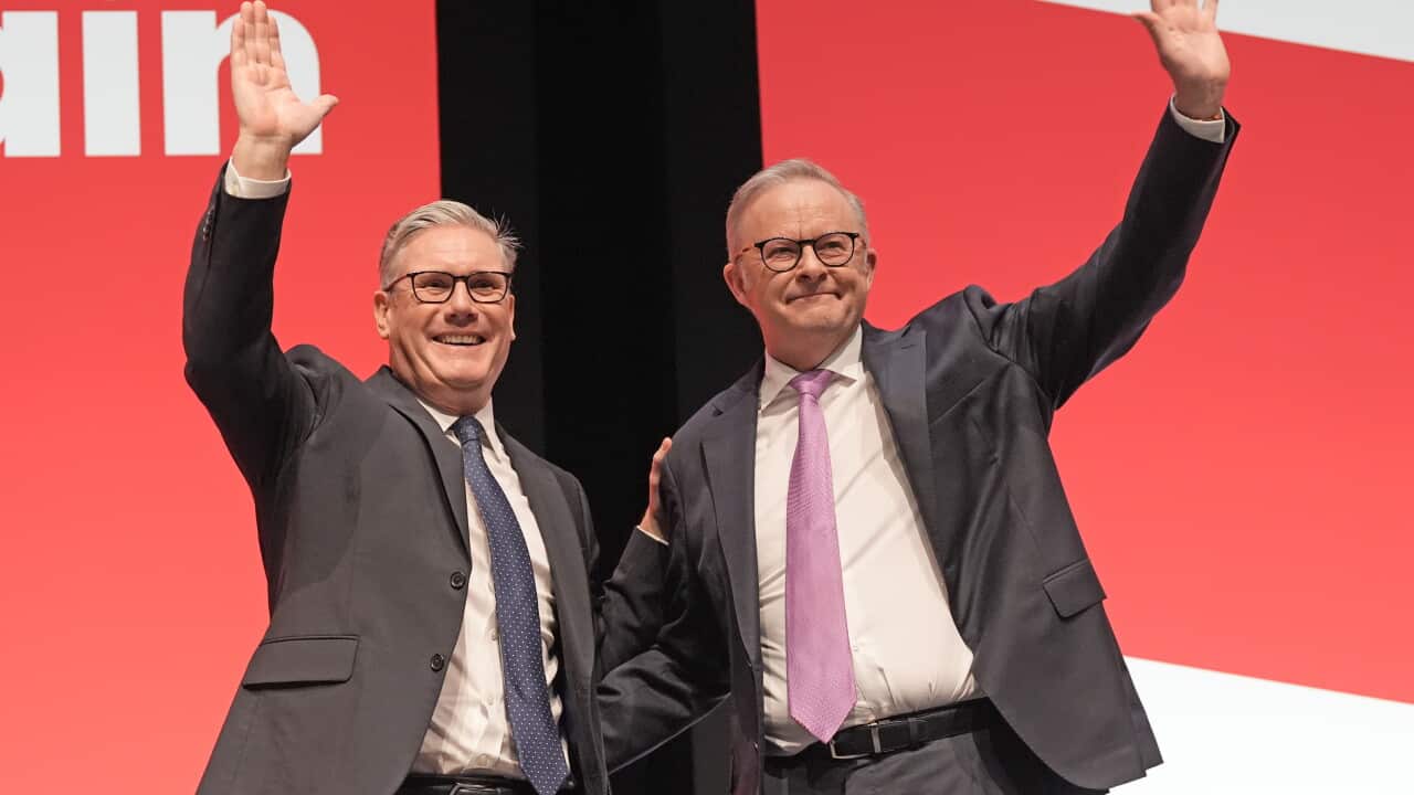 Two men in suits waving on stage against a red background.