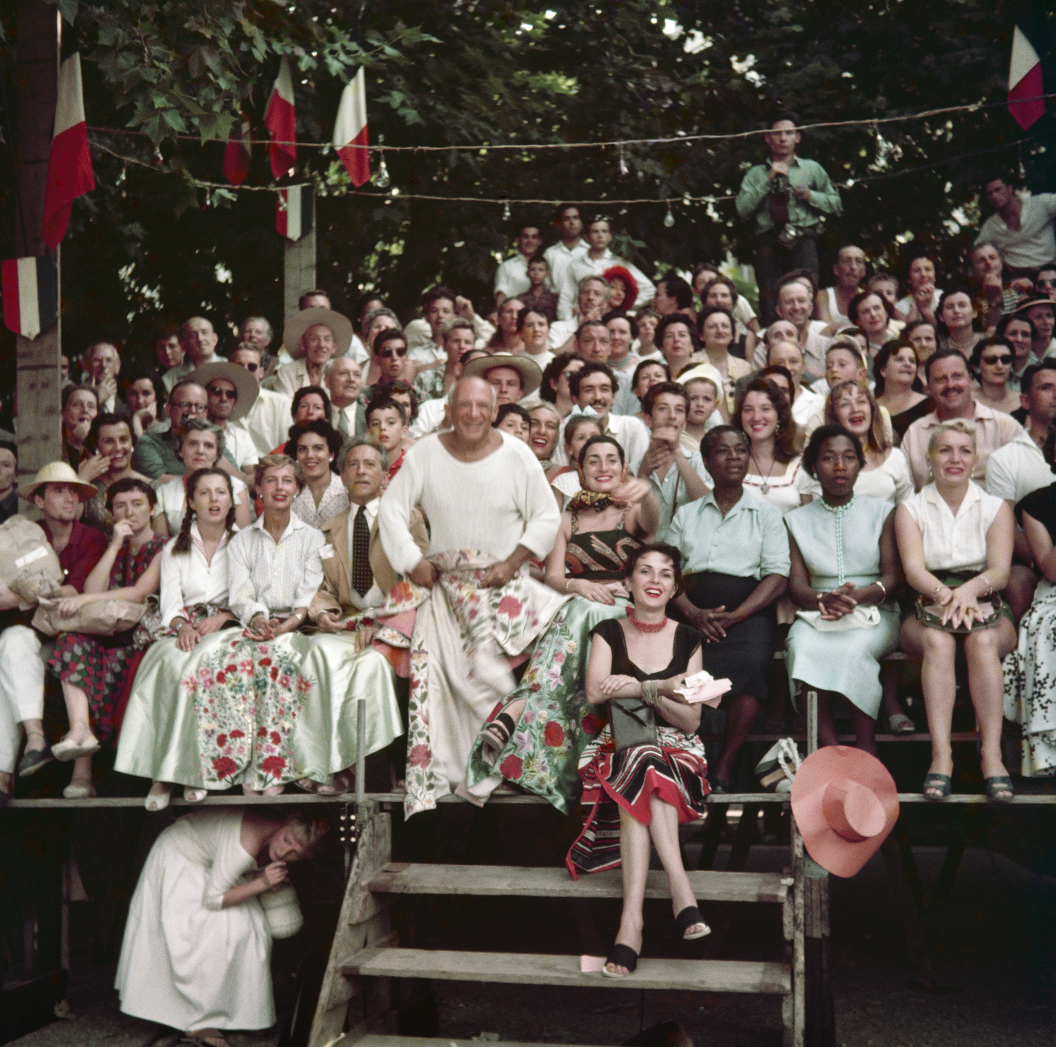 Pablo Picasso stands smiling among a packed, sunlit crowd of spectators decked with French tricolour flags and bunting.