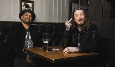 CJ Wildheart and Ginger Wildheart sitting in a pub, with Ginger extending a middle finger