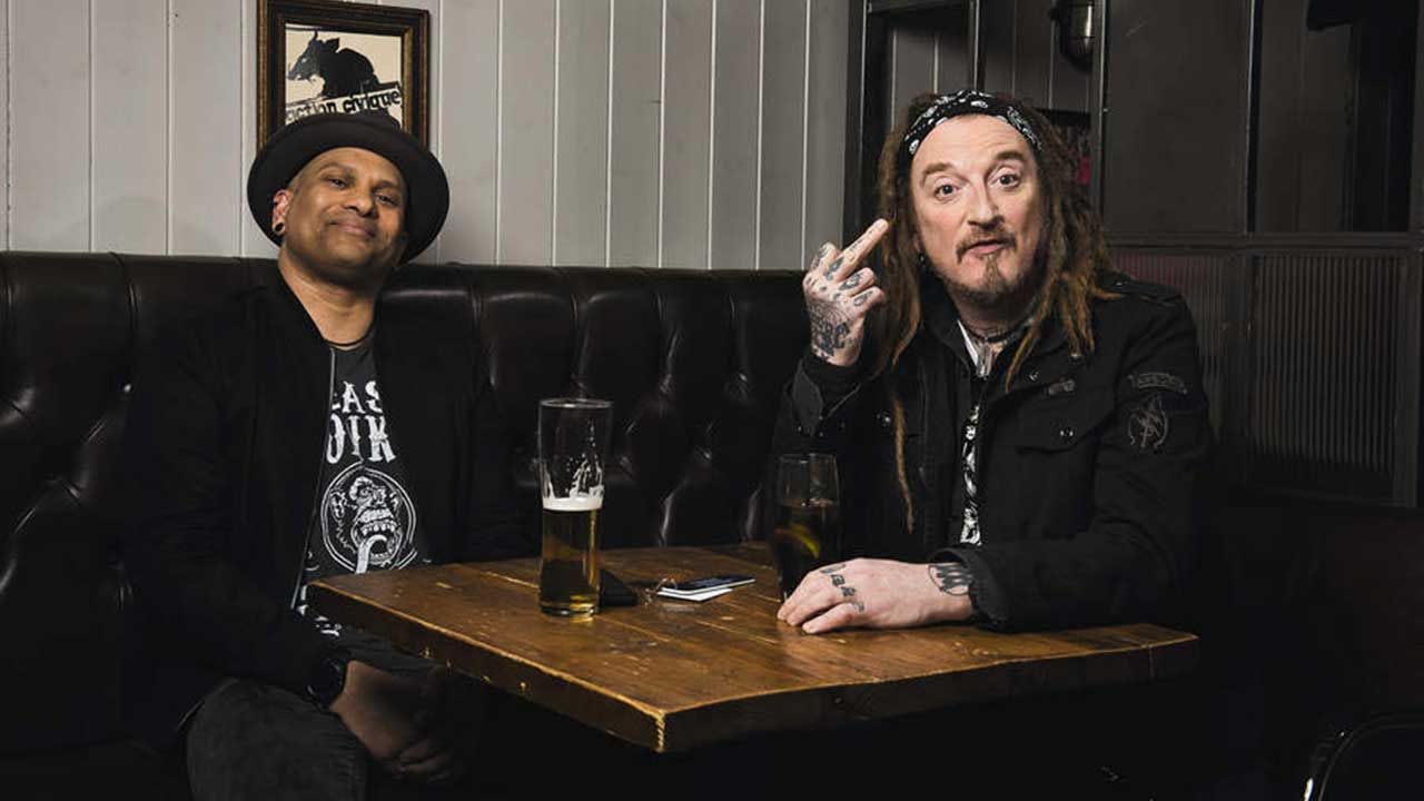 CJ Wildheart and Ginger Wildheart sitting in a pub, with Ginger extending a middle finger
