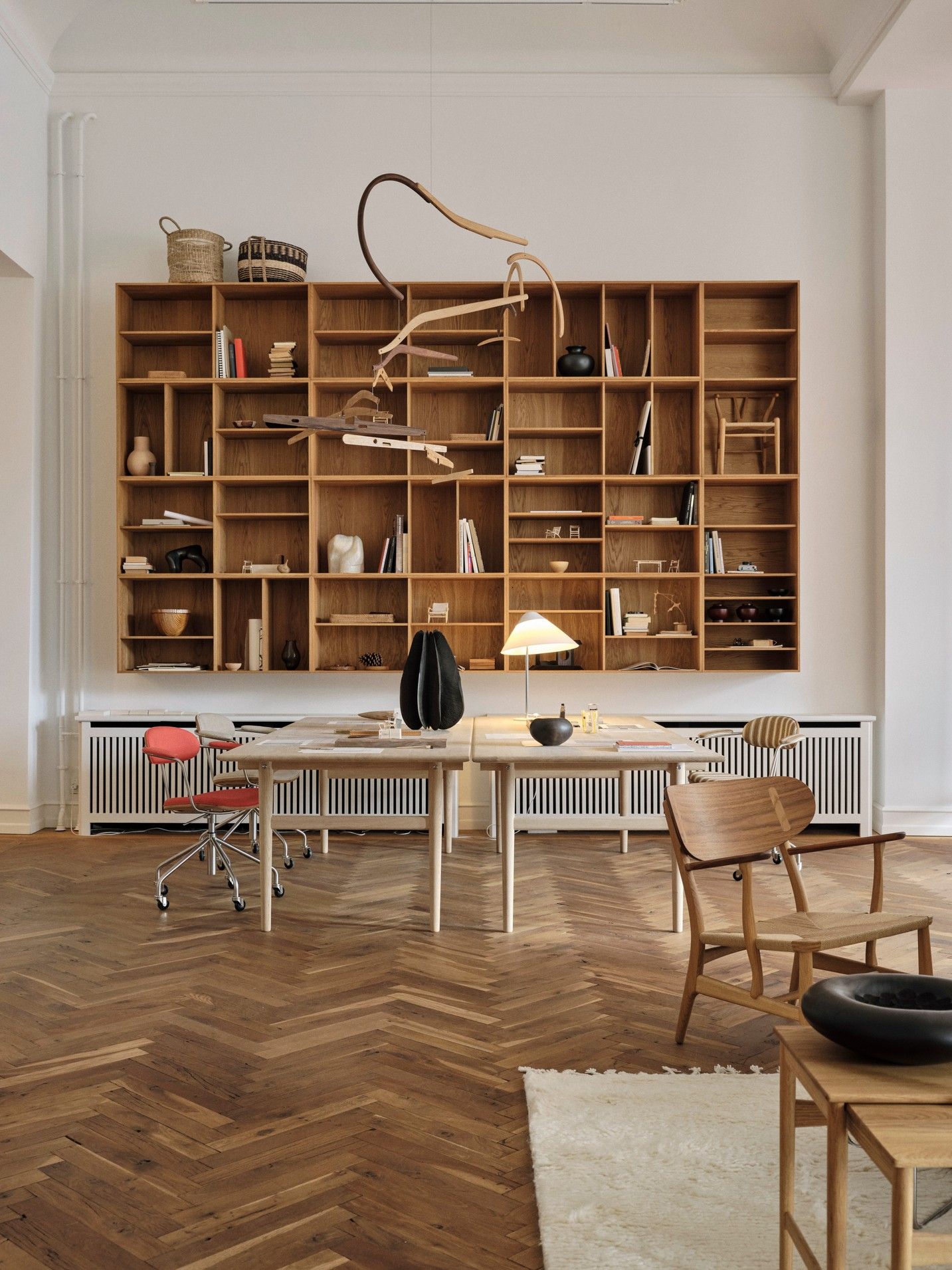 A wood-clad design showroom boasts a large, wall-hung bookcase filled with books, design prototypes, and collectibles, a pale wood desk with industrial chrome and fabric chairs, and wooden floors.