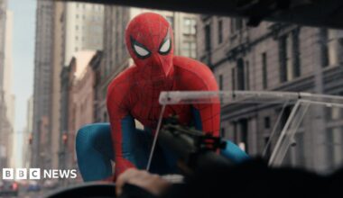Spiderman on the bonnet of a car, with a number of tall stone buildings behind him.