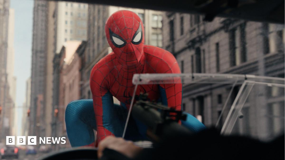 Spiderman on the bonnet of a car, with a number of tall stone buildings behind him.