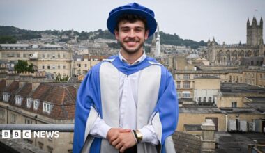 George Clarke is standing on a balcony overlooking the city of Bath. He is dressed in a graduation cap and gown after being given an Honorary Doctorate of Arts. The gown and cap are blue and silver. George has short dark hair and facial hair and is standing with his hands clasped in front of him, looking at the camera and smiling.