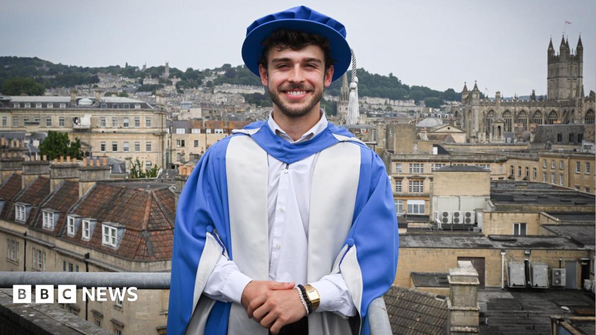 George Clarke is standing on a balcony overlooking the city of Bath. He is dressed in a graduation cap and gown after being given an Honorary Doctorate of Arts. The gown and cap are blue and silver. George has short dark hair and facial hair and is standing with his hands clasped in front of him, looking at the camera and smiling.