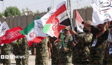 Iranian military personnel stand in a line waving red, white, green and black flags.