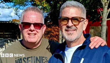 Mike, left, has short grey hair and is smiling at the camera. He is wearing red tint sunglasses and a khaki green t-shirt. He has his arm around Terry's shoulder. Terry, right, has grey hair and a grey beard. he is wearing brown sunglasses and a blue zip-up hoodie.