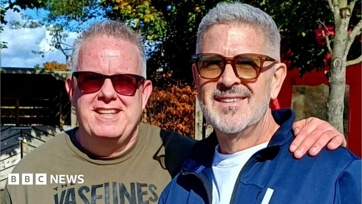 Mike, left, has short grey hair and is smiling at the camera. He is wearing red tint sunglasses and a khaki green t-shirt. He has his arm around Terry's shoulder. Terry, right, has grey hair and a grey beard. he is wearing brown sunglasses and a blue zip-up hoodie.