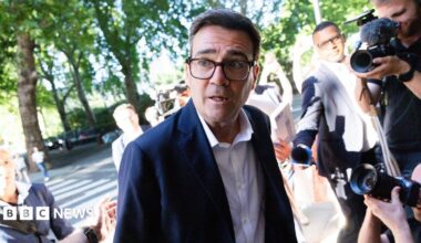Andy Burnham looks to the left of frame while wearing a suit and shirt and surrounded by cameras and reporters, as he arrives at Millbank on 2 July