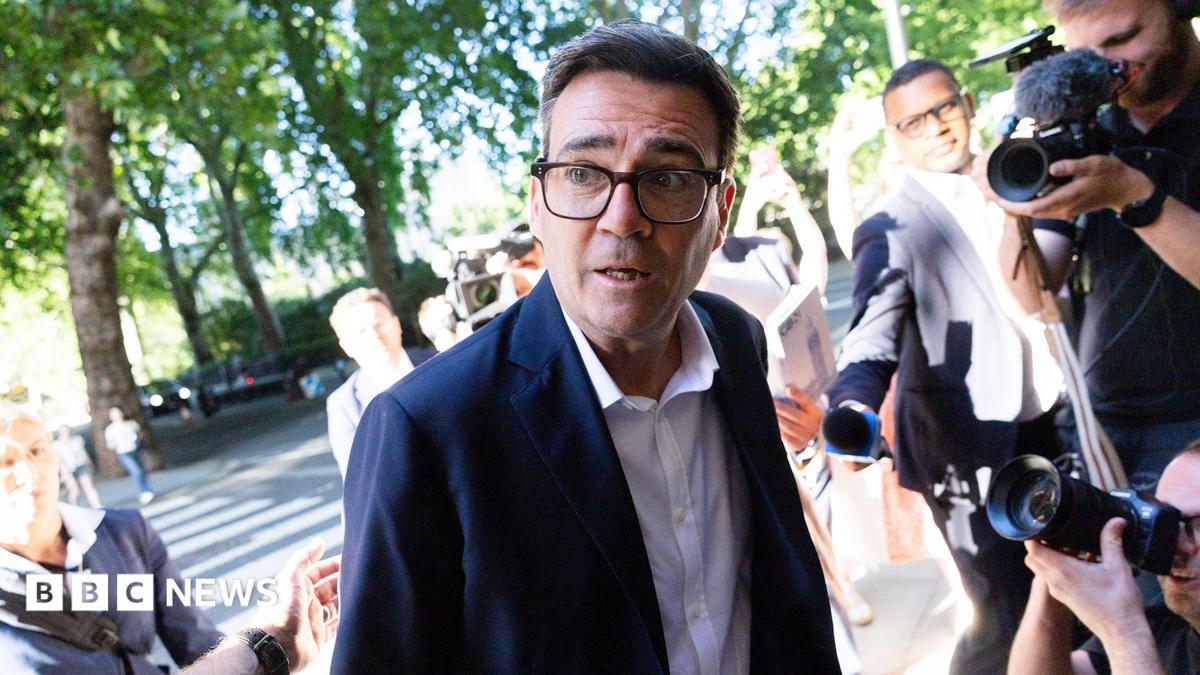 Andy Burnham looks to the left of frame while wearing a suit and shirt and surrounded by cameras and reporters, as he arrives at Millbank on 2 July