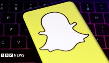 A closeup of Snapchat's logo, a white cartoon ghost with a bright yellow background, held over a laptop conputer keyboard.