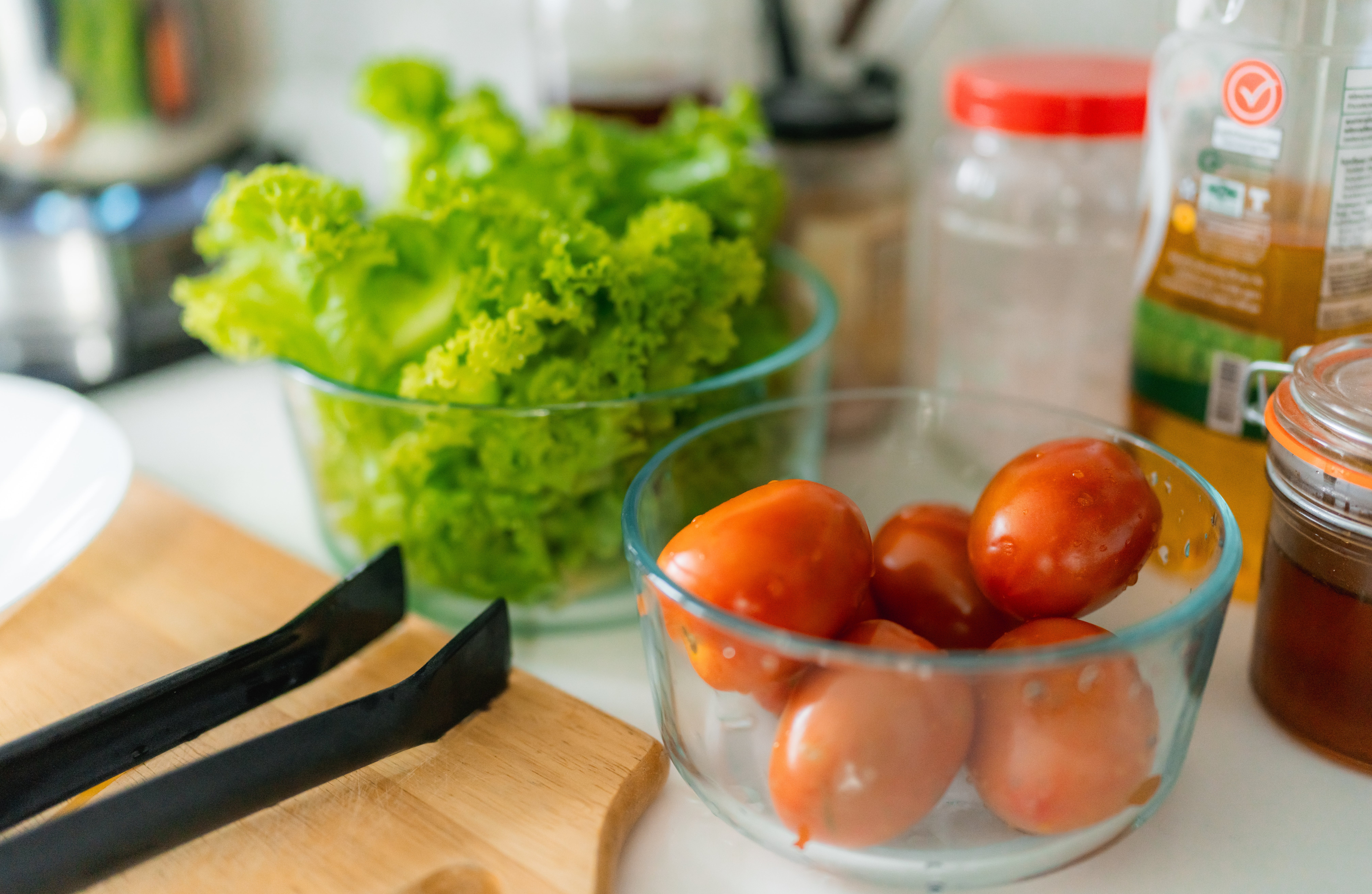 Kitchen countertop with fresh lettuce and tomatoes in glass bowls, surrounded by bottles and jars, indicating meal preparation or cooking