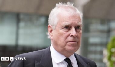 Andrew Mountbatten-Windsor is seen in a black suit and tie with a white shirt outside a building. The background is out of focus but appears to show him outside.