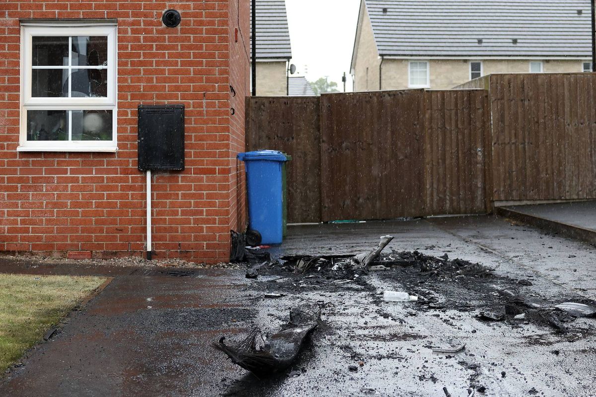 An outdoor area featuring a brick building with glass windows and a black object attached to the wall. A blue trash bin is situated near a wooden fence. The ground is covered in wet debris, suggesting a recent incident or spill.