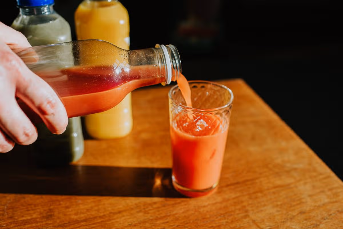 A hand holds a plastic bottle of red berry juice and pours the liquid into a glass. Two other bottles of delicious juice sit off to the side.
