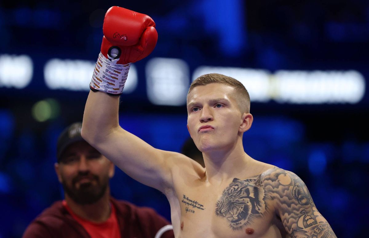 An individual with tattoos on his arms raises his left hand while wearing red boxing gloves. A person stands behind him with his back turned, partially visible against a blurred blue background.