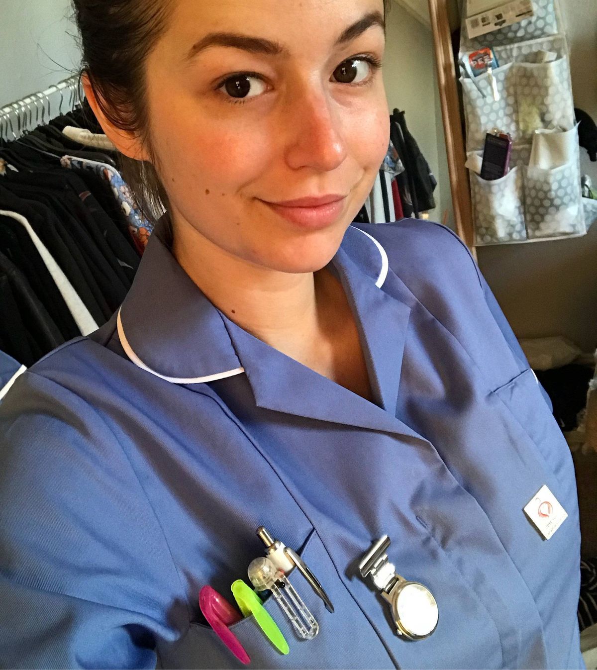 A healthcare professional in a blue uniform with a white collar, wearing various medical tools on their chest and smiling at the camera.
