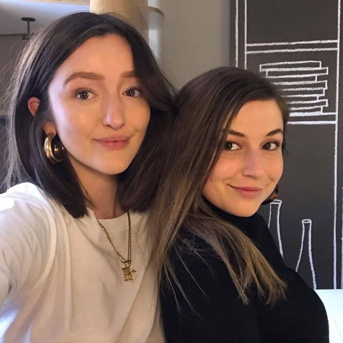 Two individuals, one wearing a white top and the other a black top, are standing together and smiling for a photograph. They are in front of a backdrop with a geometric pattern.