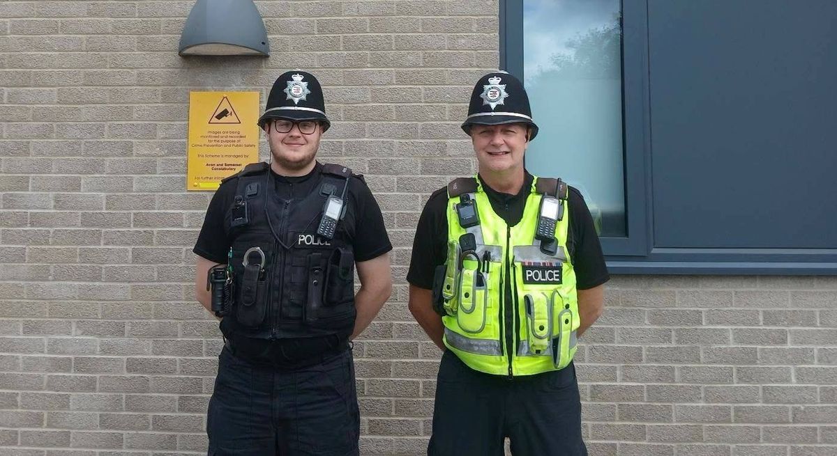 Two individuals dressed in police uniforms are standing side by side in front of a brick wall. One is wearing a black uniform and a black helmet, while the other is wearing a yellow vest over a black uniform and a black helmet.