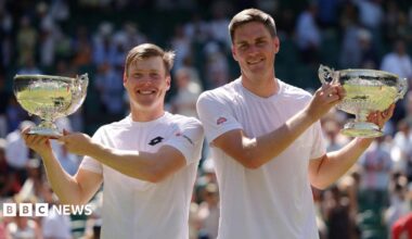 Harri Heliovaara and Henry Patten holding their large trophies aloft on Wimbledon's Centre Court. They are both wearing white T-shirts and smiling.
