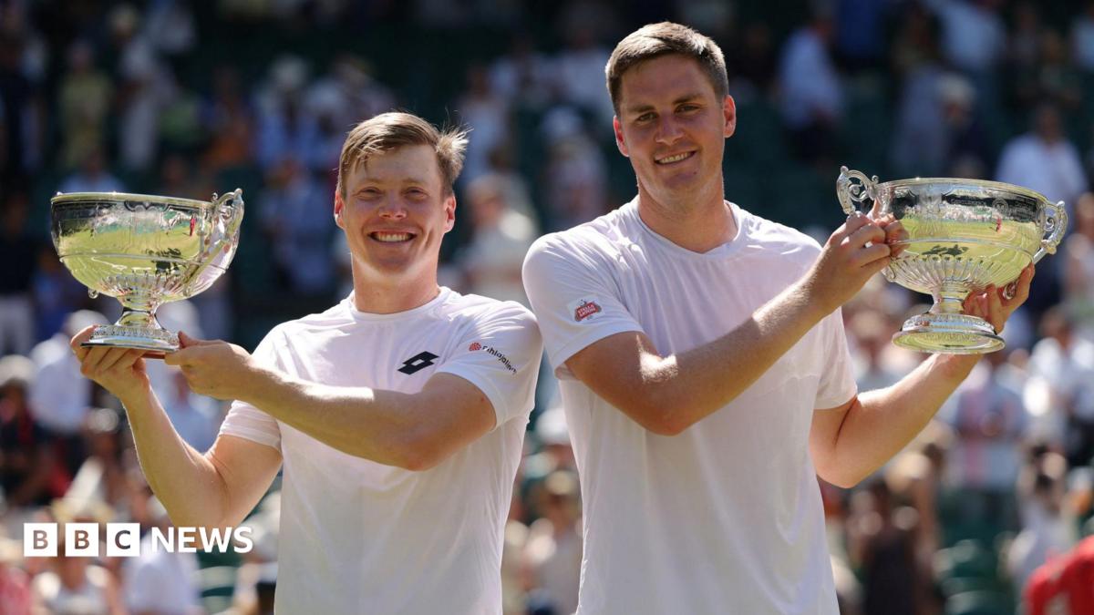 Harri Heliovaara and Henry Patten holding their large trophies aloft on Wimbledon's Centre Court. They are both wearing white T-shirts and smiling.