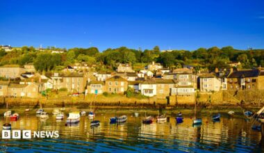 A picture of Mousehole. There are houses on the harbour. In front of the houses are small boats moored in the water. The sky is blue.