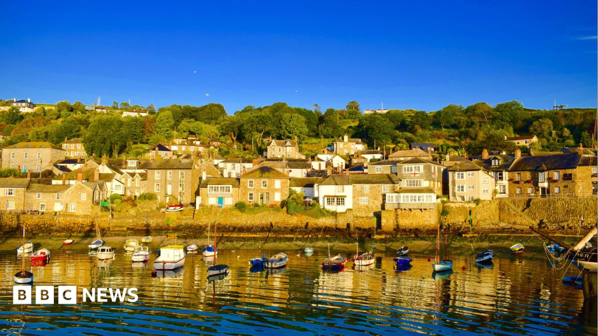 A picture of Mousehole. There are houses on the harbour. In front of the houses are small boats moored in the water. The sky is blue.
