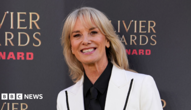 Tamzin Outhwaite smiling for the cameras in a white blazer. She is stood beside a photo board with the logo of the Olivier Awards.