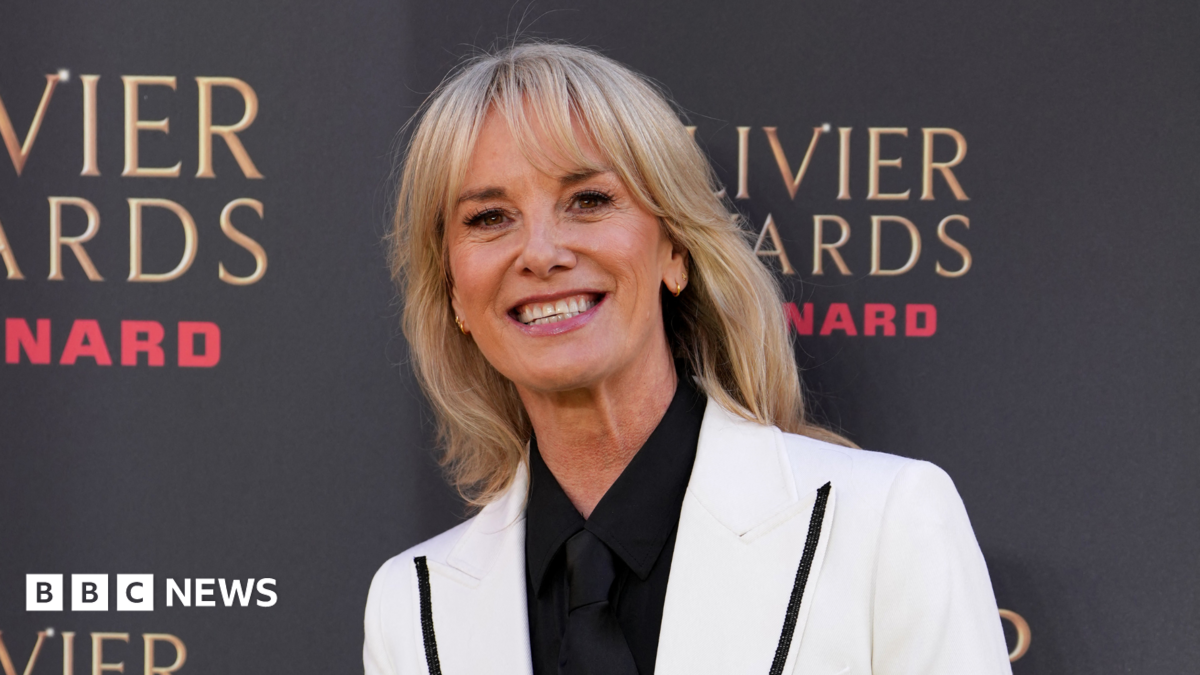 Tamzin Outhwaite smiling for the cameras in a white blazer. She is stood beside a photo board with the logo of the Olivier Awards.