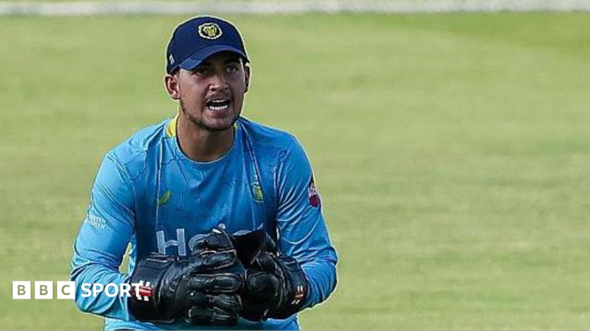 Kai Smith in action for Warwickshire in a T20 Blast game. He is wearing a light blue Bears top, navy trousers and wicket-keeper gloves and pads.