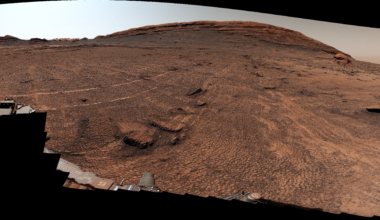 A panoramic view of a sweeping Martian plain covered in patterned fractures leading toward distant hills showcases the terrain explored by NASA's Curiosity rover to investigate the planet's past environment.