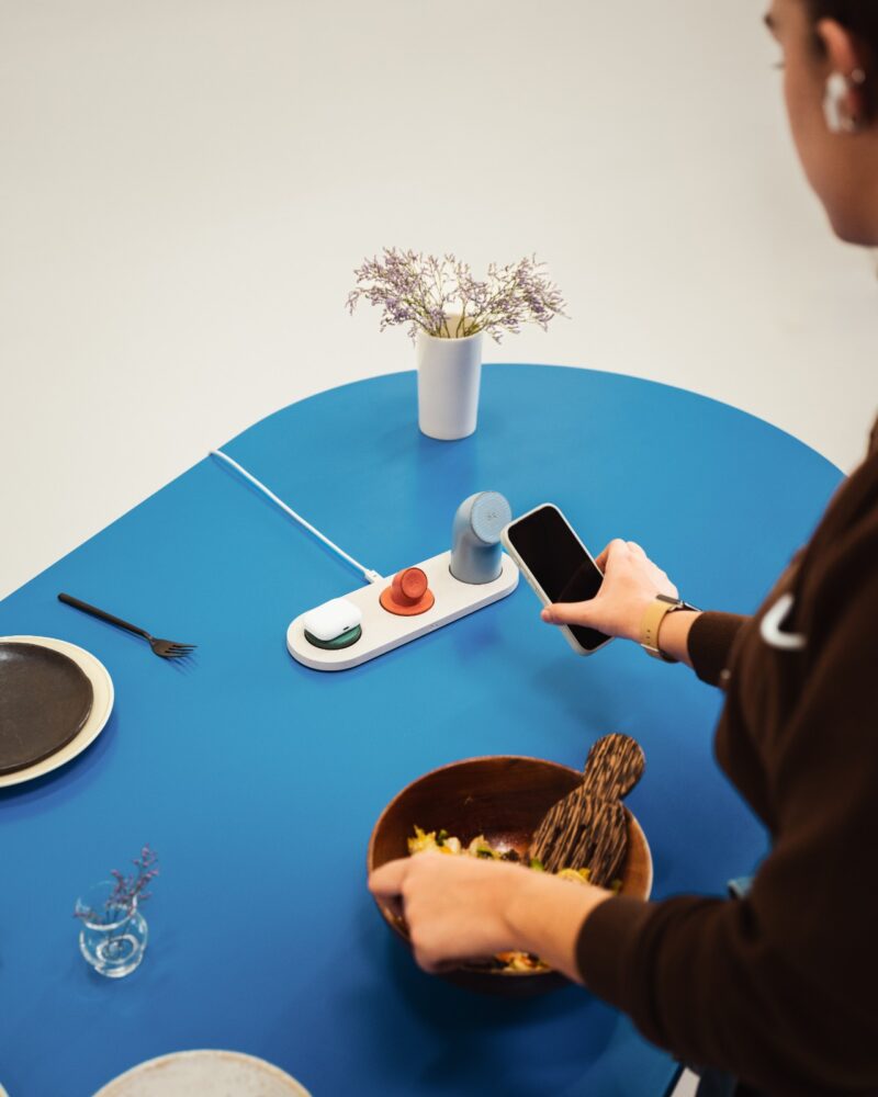 Person standing at a blue table preparing food, holding a phone near a DailyObjects wireless charging station, with plates, utensils, and a vase with flowers on the table.