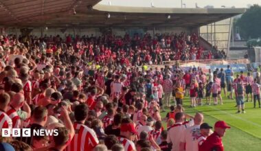 Fans in an L-shaped stand face off. Most wear red and white. Some fans are on the pitch.