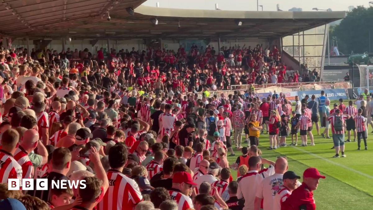 Fans in an L-shaped stand face off. Most wear red and white. Some fans are on the pitch.