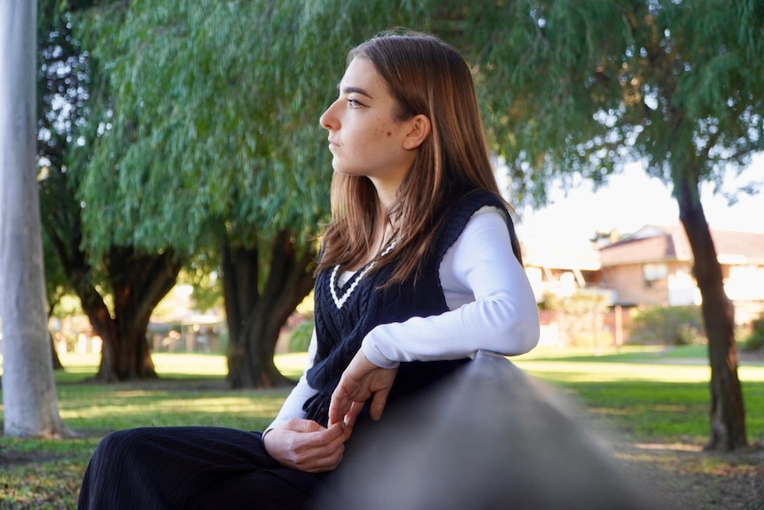 A young woman sits on a bench in a park looking away. 