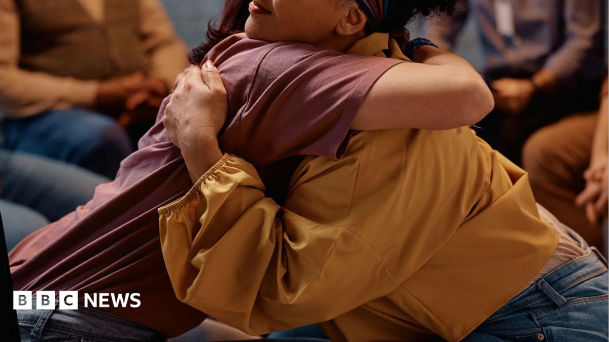 Two people sat on chairs in a group lean into each other for a hug. The person on the left is wearing a maroon top while the person on the right is wearing a yellow blouse with puffed sleeves.