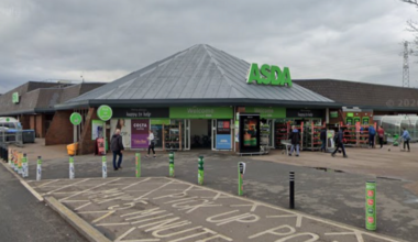 A wide view shows the entrance of an ASDA supermarket, with the green ASDA sign above the main doors and several shoppers walking near the entrance. The foreground includes a marked pick-up point area with painted road text and green bollards lining the pavement. The store is a low brick building beneath a cloudy sky, with parking areas and a few trees visible around the site.
