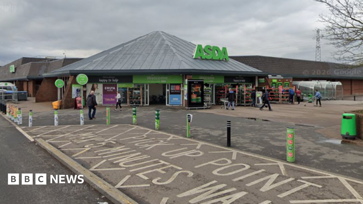 A wide view shows the entrance of an ASDA supermarket, with the green ASDA sign above the main doors and several shoppers walking near the entrance. The foreground includes a marked pick-up point area with painted road text and green bollards lining the pavement. The store is a low brick building beneath a cloudy sky, with parking areas and a few trees visible around the site.